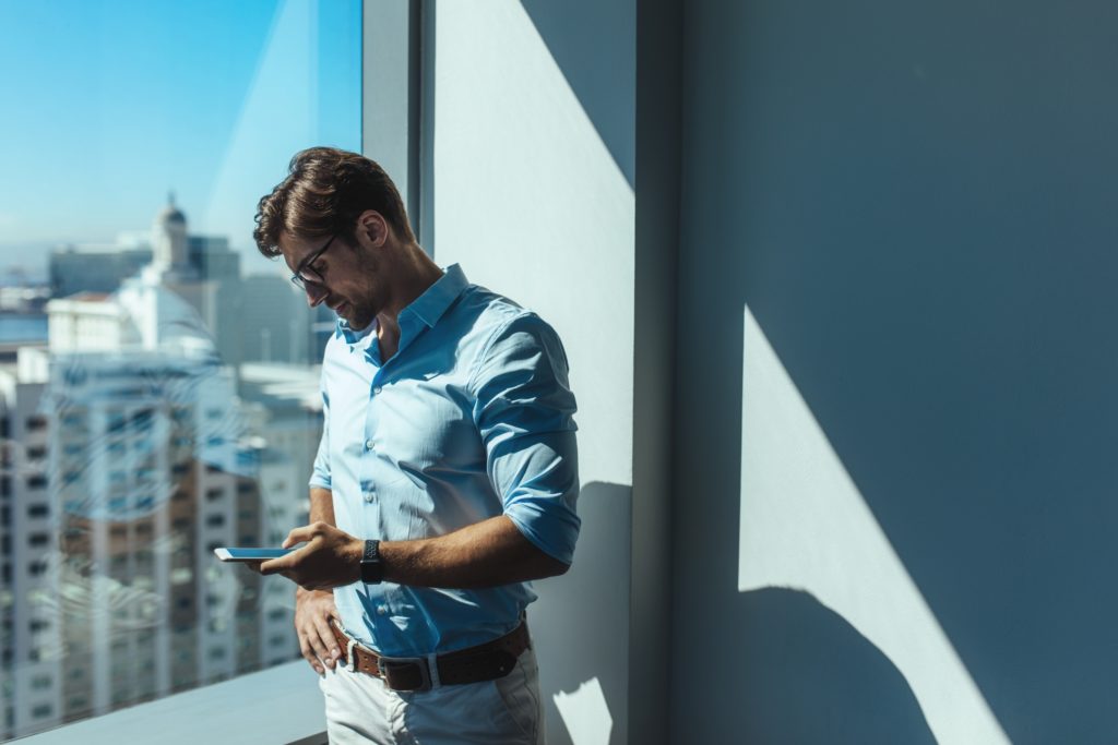 Young businessman looking at mobile phone standing beside a window. Man dressed in formals standing near window of highrise office building overlooking the cityscape.